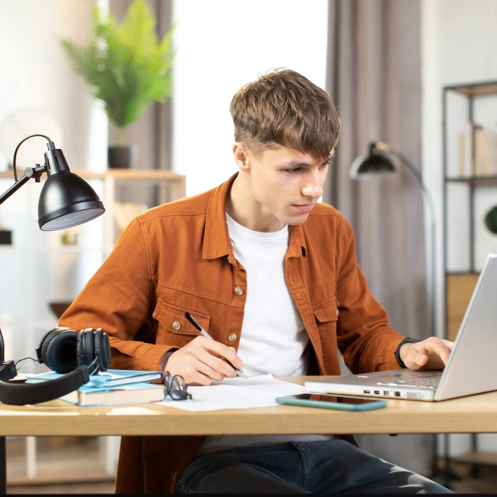 Focused young man sitting at table with laptop and making notes